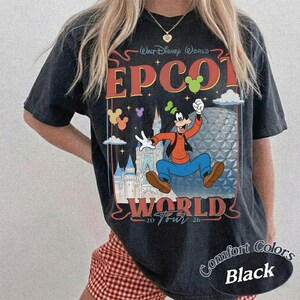 以下が含まれることがあります： Walt Disney World EPCOT WORLD 20 26というテキストがプリントされた黒いTシャツ。デザインには、グーフィーのイラスト、スペースシップ・アースの建造物、城が描かれており、小さなカラフルなミッキーのアイコンがアクセントになっています。