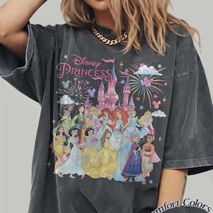May include: Dark gray t-shirt featuring a graphic of Disney Princesses in front of a castle with fireworks. The text "Disney Princess" is above the castle. The shirt is labeled "Comfort Colors Pepper".