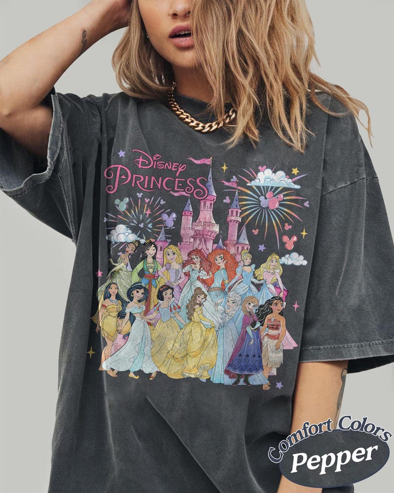 May include: Dark gray t-shirt featuring a graphic of Disney Princesses in front of a castle with fireworks. The text "Disney Princess" is above the castle. The shirt is labeled "Comfort Colors Pepper".
