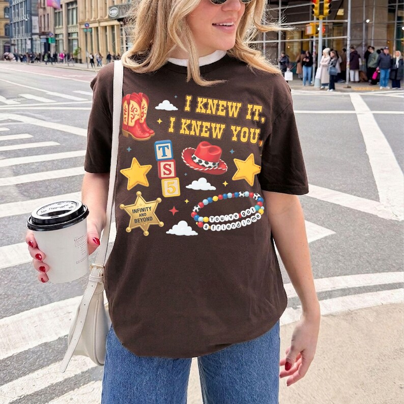 Pode incluir: Uma t-shirt de manga curta castanho-escura com uma impress&atilde;o gr&aacute;fica que inclui o texto I KNEW IT, I KNEW YOU, uma bota de cowboy vermelha, um chap&eacute;u de cowboy vermelho, estrelas amarelas, uma estrela de xerife e uma pulseira de contas.