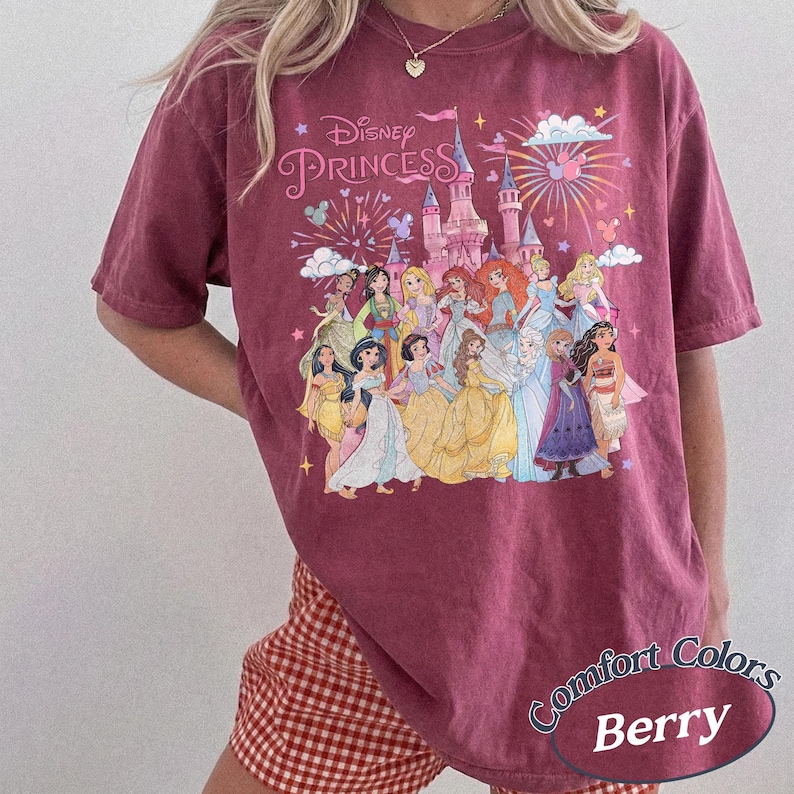 May include: Berry-colored t-shirt featuring a graphic of Disney Princesses in front of a castle with fireworks. The text "Disney Princess" is above the princesses. The shirt also has the text "Comfort Colors Berry".
