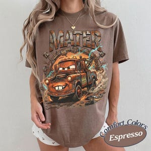 May include: A brown short-sleeve t-shirt featuring a graphic print of the character Mater from the movie Cars. The design includes the text MATER in a rusted metal font, surrounded by mechanical parts like pistons and wrenches with lightning.