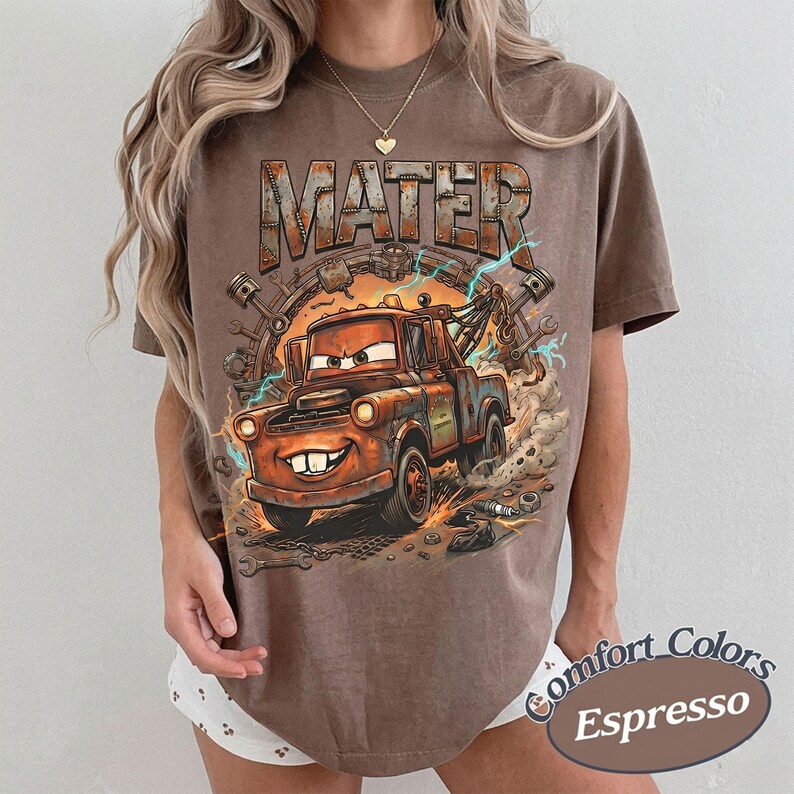 May include: A brown short-sleeve t-shirt featuring a graphic print of the character Mater from the movie Cars. The design includes the text MATER in a rusted metal font, surrounded by mechanical parts like pistons and wrenches with lightning.