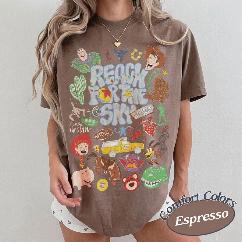Peut inclure: Un t-shirt marron &agrave; manches courtes avec une impression graphique portant le texte REACH FOR THE SKY. Le motif comprend divers personnages de dessins anim&eacute;s et des ic&ocirc;nes de film, tels qu'un cow-boy, un ranger de l'espace et une voiture jaune.
