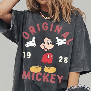 Op de afbeelding: Een antracietgrijs T-shirt met korte mouwen en een afbeelding van Mickey Mouse met de tekst ORIGINAL MICKEY 19 28. Het shirt heeft een vervaagde uitstraling en bevat een klein rond logo in de hoek met de tekst Comfort Colors Pepper.