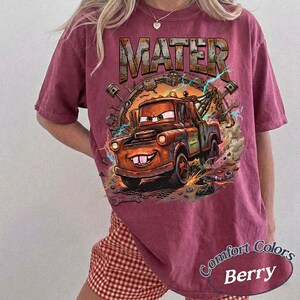 May include: A berry-colored short-sleeve t-shirt featuring a graphic print of a rusty brown tow truck character with large eyes and buck teeth. The text MATER is printed in a metallic-style font above the truck, surrounded by mechanical tools.