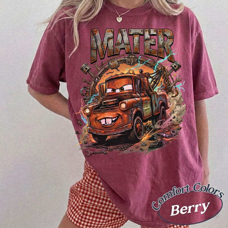May include: A berry-colored short-sleeve t-shirt featuring a graphic print of a rusty brown tow truck character with large eyes and buck teeth. The text MATER is printed in a metallic-style font above the truck, surrounded by mechanical tools.