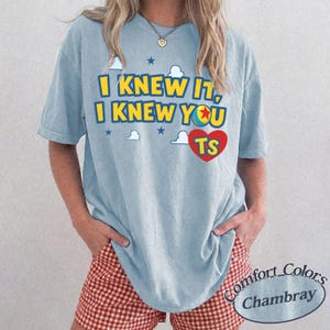 K&ouml;nnte beinhalten: Ein hellblaues Kurzarm-T-Shirt mit dem gelben Text I KNEW IT, I KNEW YOU, einer Ballgrafik anstelle des Buchstabens O und einem roten Herz mit den Buchstaben TS, umgeben von kleinen wei&szlig;en Wolken und blauen Sternen.