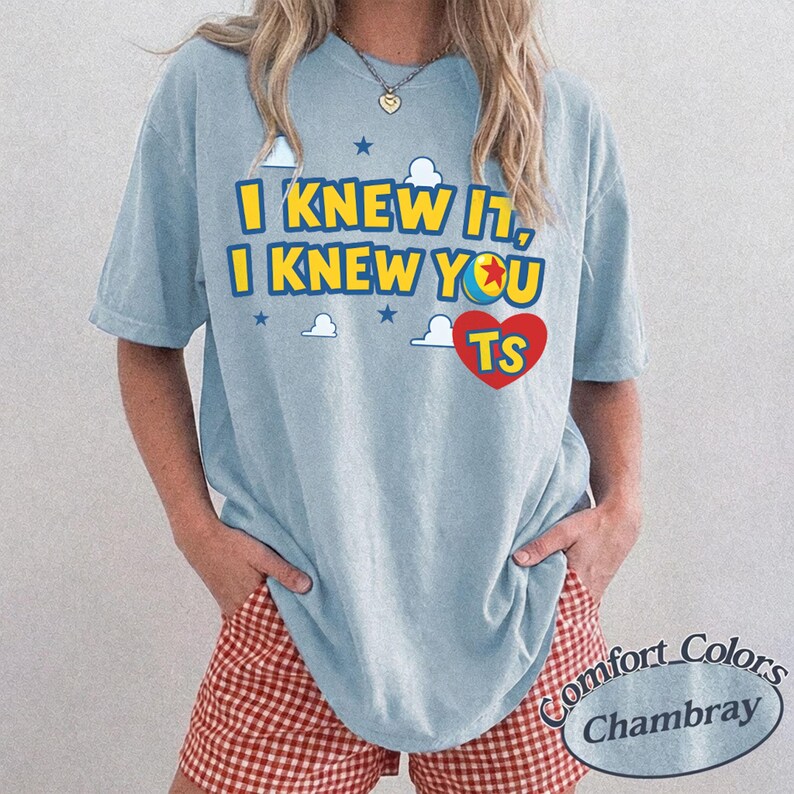 K&ouml;nnte beinhalten: Ein hellblaues Kurzarm-T-Shirt mit dem gelben Text I KNEW IT, I KNEW YOU, einer Ballgrafik anstelle des Buchstabens O und einem roten Herz mit den Buchstaben TS, umgeben von kleinen wei&szlig;en Wolken und blauen Sternen.