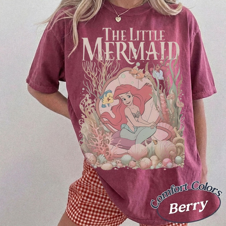 Pu&ograve; includere: Una t-shirt a maniche corte color bacca con una stampa grafica di The Little Mermaid e il testo The Little Mermaid sopra una scena che ritrae una sirena seduta in una grande conchiglia circondata da coralli, alghe e varie conchiglie.