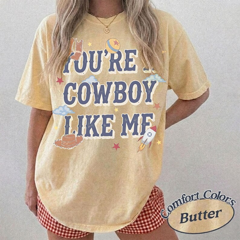 Op de afbeelding: Een geel t-shirt met korte mouwen en een verwassen textuur. Op de voorkant staat de donkerblauwe tekst "YOU'RE A COWBOY LIKE ME", omringd door grafische illustraties van een cowboylaars, een hoed, een raket, sterren, wolken en een bal.