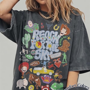Peut inclure: Un t-shirt gris anthracite arborant une impression graphique color&eacute;e avec le texte "REACH FOR THE SKY" entour&eacute; de divers personnages de dessins anim&eacute;s et d'ic&ocirc;nes, dont un cow-boy, un ranger de l'espace, un camion jaune et d'autres illustrations.