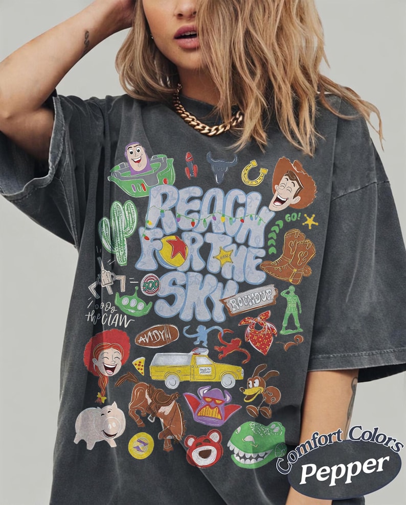 Peut inclure: Un t-shirt gris anthracite arborant une impression graphique color&eacute;e avec le texte "REACH FOR THE SKY" entour&eacute; de divers personnages de dessins anim&eacute;s et d'ic&ocirc;nes, dont un cow-boy, un ranger de l'espace, un camion jaune et d'autres illustrations.