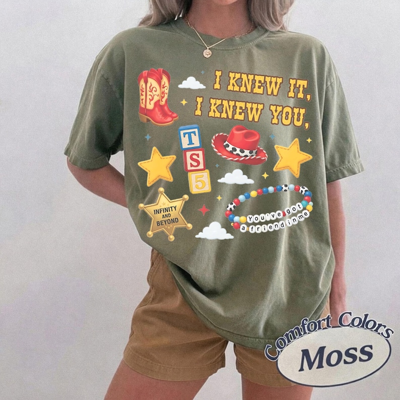 Pode incluir: Uma t-shirt verde-oliva com uma estampa gr&aacute;fica contendo o texto I KNEW IT, I KNEW YOU, uma bota de cowboy vermelha, um chap&eacute;u de cowboy vermelho, estrelas amarelas, uma estrela de xerife com o texto INFINITY AND BEYOND, blocos e uma pulseira.