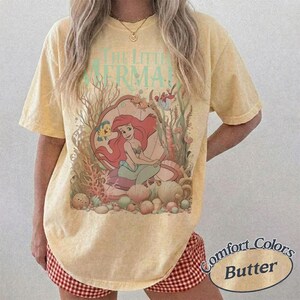 Pu&ograve; includere: Una t-shirt a maniche corte color giallo pallido con una grafica del personaggio Ariel de La Sirenetta seduta all'interno di una grande conchiglia circondata da coralli e vita marina. Il testo The Little Mermaid &egrave; stampato in turchese chiaro.