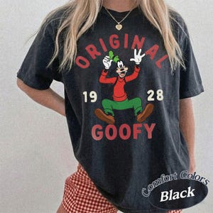 Op de afbeelding: Een zwart t-shirt met korte mouwen en een afbeelding van het stripfiguur Goofy. De tekst luidt ORIGINAL GOOFY 19 28. Goofy draagt een rood shirt met lange mouwen, een groene broek en bruine schoenen, en houdt een groene hoed in de hand.
