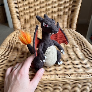 May include: A dark brown crocheted dragon figure with a cream-colored belly, red inner wings, and a bright orange and yellow flame at the tip of its tail. The creature has sharp white claws, pointed ears, and red eyes, crafted in a yarn-based style.