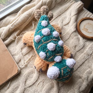 May include: A plush crochet dinosaur toy featuring a soft peach-coloured body with a teal back and head. The creature is adorned with white pom-poms along its spine and head, and it has small dark brown accents on its feet and tail tip.