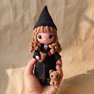 May include: A crocheted doll featuring a character with light brown curly hair, wearing a black pointed hat and a black robe with a red and gold emblem. The doll holds a small brown book and stands next to a tiny brown crocheted cat figure.