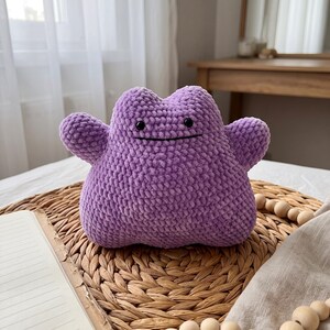 May include: A purple plush toy with a rounded, blob-like body and two small arms extending from the sides. The toy features a textured, yarn-like surface, two small black circular eyes, and a simple, thin black horizontal line for a mouth.