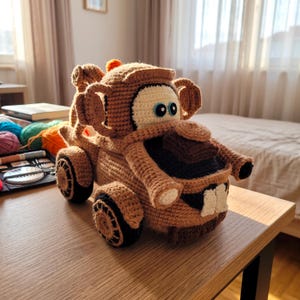 Pode incluir: Um caminhão de brinquedo de crochê marrom com grandes olhos brancos de pupilas azuis, uma grade frontal preta e dois dentes dianteiros brancos proeminentes. O veículo possui um gancho de reboque na traseira, rodas texturizadas e um pequeno detalhe laranja no teto.
