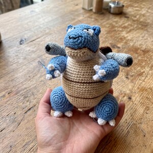 May include: A crocheted stuffed toy shaped like a blue turtle creature with a tan belly, dark brown shell, and two gray cannons on its back. The figure features white claws on its hands and feet, a stitched mouth, and white accents around the eyes.