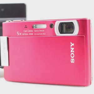 May include: A bright pink Sony digital camera with a Carl Zeiss Vario-Tessar lens and 5x optical zoom. The camera has a silver accent and the Sony logo in white. A black charger and blue box are in the background.