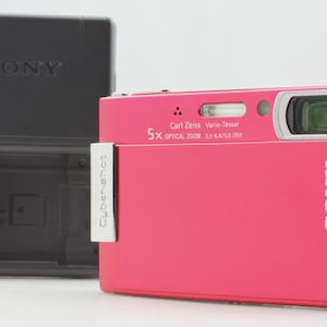 May include: A bright pink Sony Cyber-shot digital camera with a Carl Zeiss Vario-Tessar lens and 5x optical zoom, positioned next to a black Sony battery charger. The camera features a metallic silver side accent and clear branding on the front.