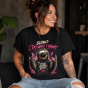 May include: A black t-shirt featuring a graphic of a sloth making hand gestures. The text reads "SLOW? I DO WHAT I WANT" in pink and "ZERO RUSHES GIVEN" in white, with a small anarchy symbol below. The sloth wears a black shirt and has pink lightning.
