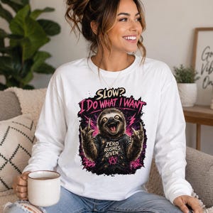 May include: A white long-sleeve shirt featuring a graphic of a sloth making hand gestures. The text reads SLOW? I DO WHAT I WANT and ZERO RUSHES GIVEN, accompanied by a pink lightning bolt design and an anarchy symbol on the sloth's black t-shirt.