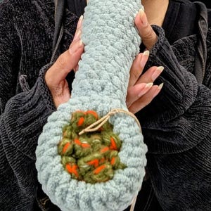 May include: A light blue, plush, crochet pipe with a round bowl filled with green and orange yarn. The pipe is held by a person wearing a black jacket.
