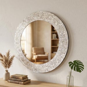 Puede incluir: Un espejo redondo decorativo con un marco blanco con motivos florales cuelga de una pared color crema. Una mesa de madera exhibe libros, un jarrón con flores secas y una hoja de monstera en un jarrón de cristal. La habitación tiene una paleta de colores neutros.