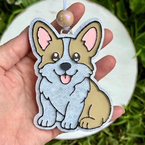 May include: A Corgi-shaped hanging ornament featuring a tan and light gray textured finish with black outlines. The dog has pink inner ears, a pink tongue, and large dark eyes. A white string with a single iridescent bead is attached.