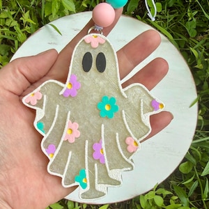 May include: A translucent, glittery ghost-shaped ornament featuring black oval eyes and colorful floral accents in pink, purple, and teal. The ghost is attached to a silver metal loop with a string of round pink and teal beads at the top.