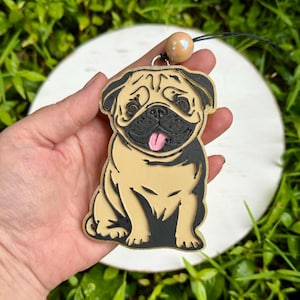 May include: A tan and black pug pendant featuring a detailed engraved design of a sitting dog with a pink tongue. The pendant is attached to a black cord with a single iridescent, round, peach-colored bead positioned above the dog figure.
