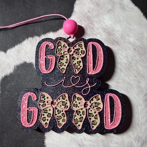 May include: A dark blue glittery hanging ornament featuring the text GOD IS GOOD in pink glittery letters. The letters O are replaced by leopard print bows. A pink bead is attached to the top with a pink string for hanging purposes.