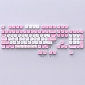 May include: A set of pink and white keyboard keycaps displayed on a light purple surface. The keycaps are in various sizes and shapes, designed for a mechanical keyboard. Some keys have decorative patterns and text. The keycaps are in various sizes and shapes.
