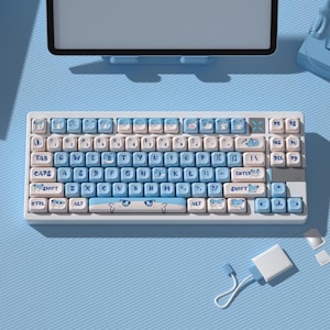 May include: A mechanical keyboard with a white frame and light blue keycaps featuring cartoon character illustrations. The spacebar displays a pair of eyes, and the keys include standard QWERTY lettering alongside various blue graphic icons.