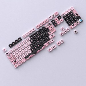 May include: A disassembled mechanical keyboard with pink and black keycaps. The keycaps feature white lettering and decorative icons. The keyboard's base is black with a pink wave design. Some keycaps are scattered around the keyboard.