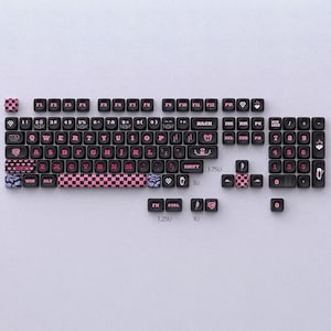 May include: Black and pink keyboard keycaps with a checkerboard pattern and heart designs. The keycaps are arranged to show a keyboard layout, with letters, numbers, and function keys visible. The keycaps are made of plastic.