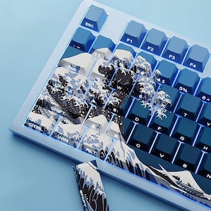 May include: A mechanical keyboard with blue keycaps and a custom wave design in white, black, and blue. The keyboard has a light blue frame and additional keycaps with the same wave pattern.