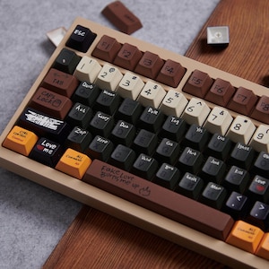 May include: A mechanical keyboard featuring a beige frame and a multi-colored keycap set in brown, black, cream, and orange. The keys display custom handwritten-style text, including phrases like "Not for Sell," "Love me," and "Fake Love Burns me up."