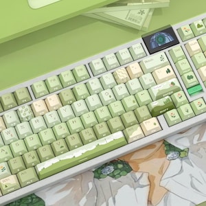 May include: A light green and white mechanical keyboard with custom keycaps. The keycaps feature a landscape design with green hills and floral accents. The keyboard has a small screen and a light green frame.