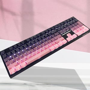 Könnte beinhalten: Eine mechanische Tastatur mit schwarzem Rahmen und einem Tastenkappen-Set im Farbverlauf von Dunkellila links bis Hellrosa rechts. Jede Tastenkappe ist mit einem weißen topografischen Kartenmuster und hintergrundbeleuchteten Zeichen versehen.