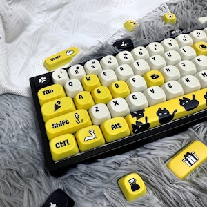 May include: A mechanical keyboard featuring a black frame and a mix of cream and yellow keycaps. The keys display black letters, numbers, and various icons including a worm, an umbrella, and cats. Additional loose yellow keycaps are scattered nearby.