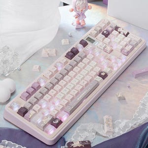 Puede incluir: Un teclado mecánico con una paleta de colores pastel en rosa claro, lavanda y blanco. Las teclas tienen impresiones gráficas personalizadas y el tablero incluye una pequeña pantalla digital. Varias teclas sueltas están esparcidas cerca.