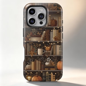 Op de afbeelding: Een telefoonhoesje met een donker, meerlaags boekenplankontwerp, versierd met herfstelementen zoals oranje pompoenen, gedroogde bladeren en kleine gloeiende lichtsnoeren. De planken staan vol met diverse boeken en een enkele witte mok.