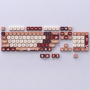 Pode incluir: Um conjunto de teclas de teclado mecânico personalizado com tema de café. As teclas são uma mistura de cores branca, castanha e bordô. As teclas têm vários ícones e texto, incluindo "RAGDOLL CAFE" e pegadas de patas de gato.