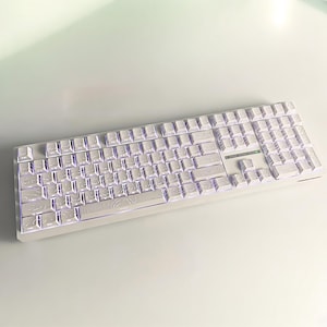 May include: A white mechanical keyboard with illuminated keys. The keyboard has a standard layout with a number pad and function keys. The keys have a textured design, and the keyboard is lit with a purple glow.