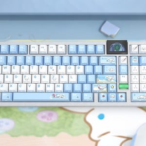 May include: A light blue and white mechanical keyboard with custom keycaps. The keycaps feature cartoon character designs and the keyboard has a small screen. The keyboard is on a light blue surface.